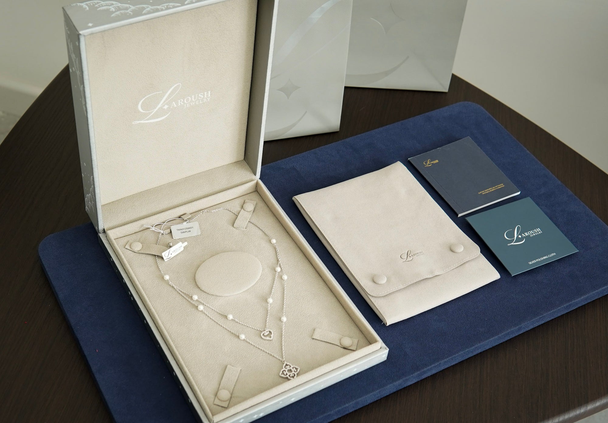 An open luxury jewelry box displaying a silver-toned necklace with pearl-like beads and a heart-shaped pendant, alongside a cream jewelry pouch, two dark blue cards, and a dark blue presentation mat on a dark wooden table.